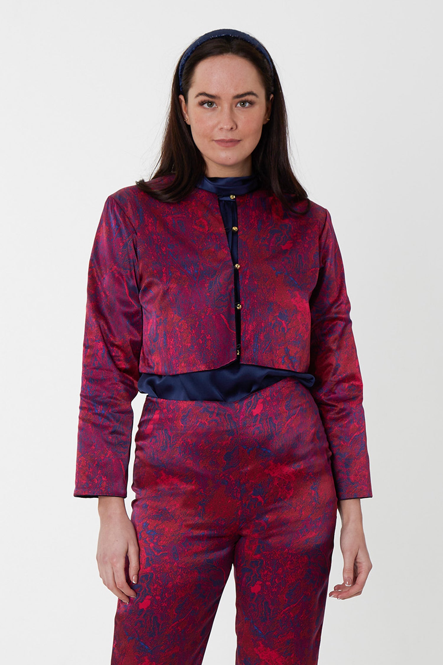 Model wearing reversible box shaped cropped red marble silk satin print jacket, with silver bell fastening, worn with matching Georgia Trousers and navy halter neck Sophie Top and headband.