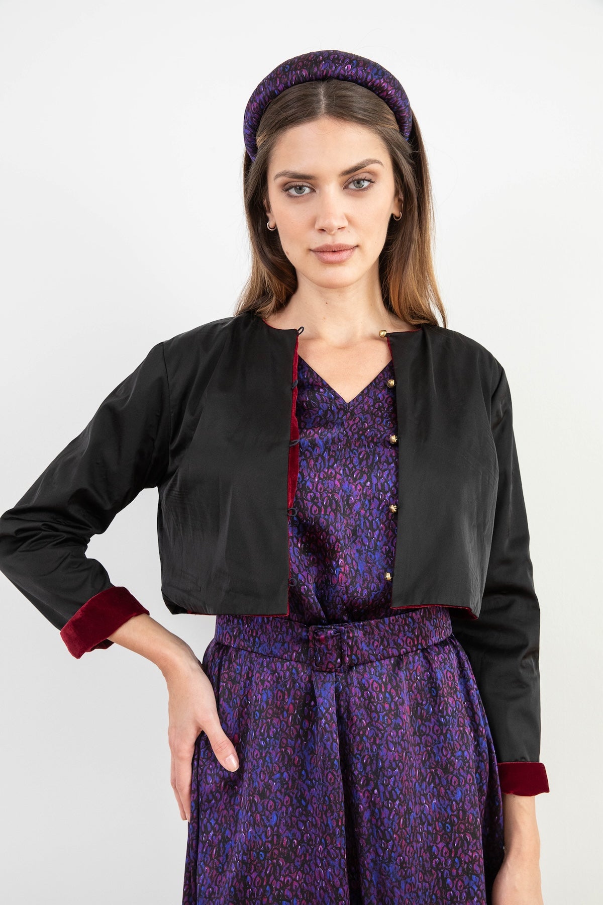 Model wearing reversible box shaped cropped black silk satin jacket, with gold bell fastening, worn with matching padded headband and purple print Valentina Dress.