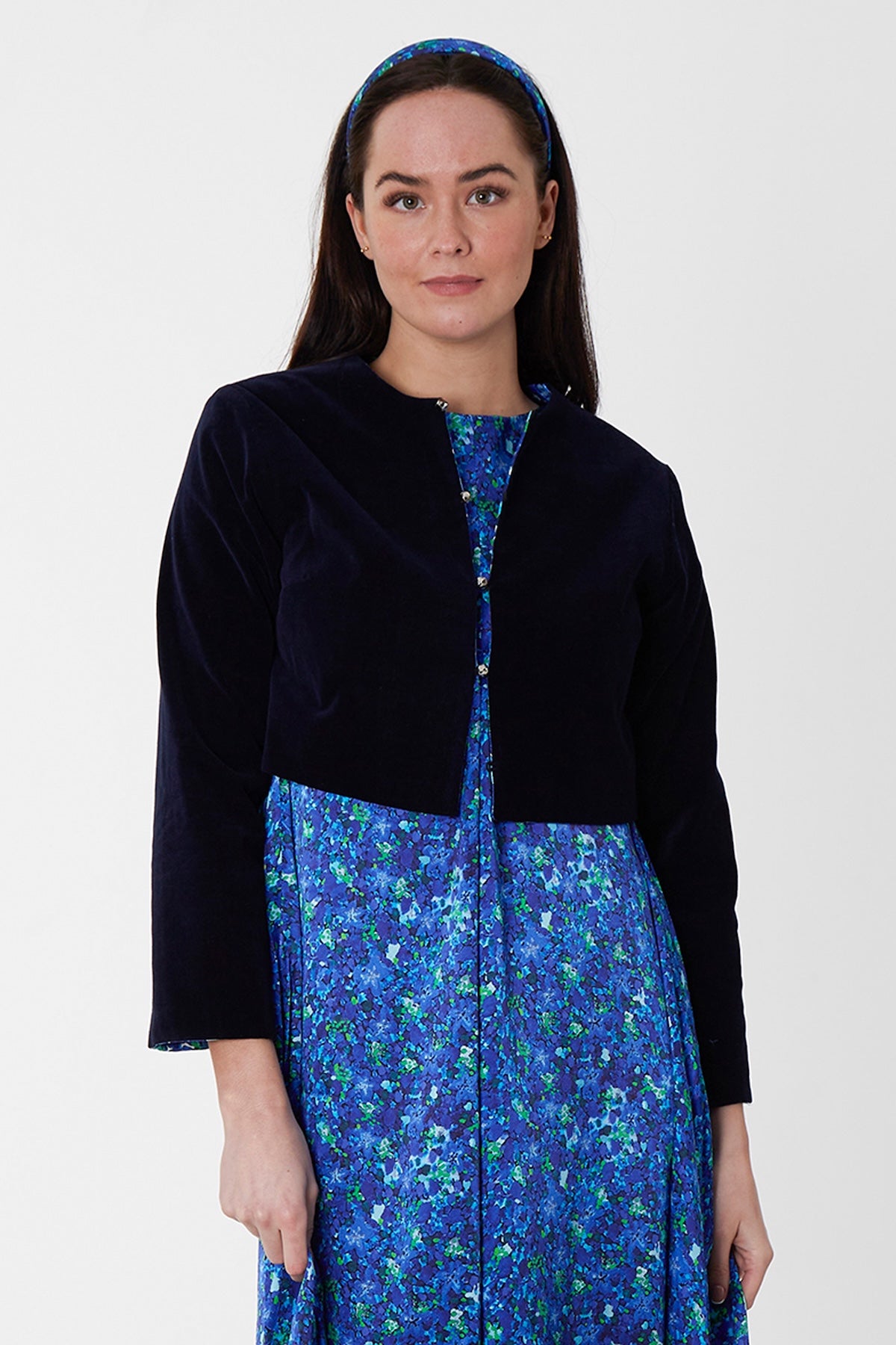 Model wearing reversible box shaped cropped navy velvet jacket, with silver bell fastening, worn with blue floral A-line Nancy Dress.