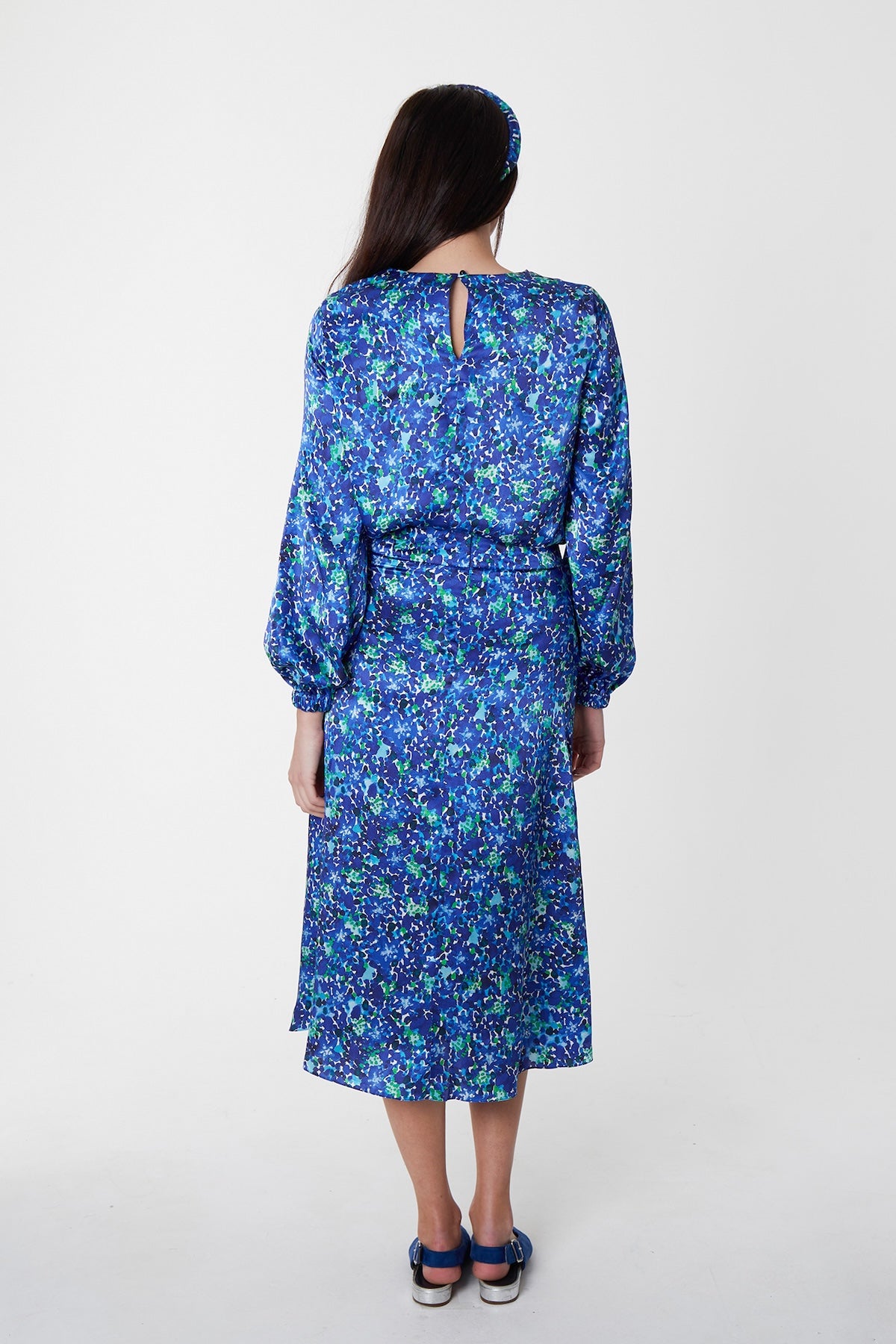 Back view of model wearing blue floral silk printed Isabel Manns narrow a-line midi skirt with wide waistband, worn with matching long sleeve Emily Top and padded headband.
