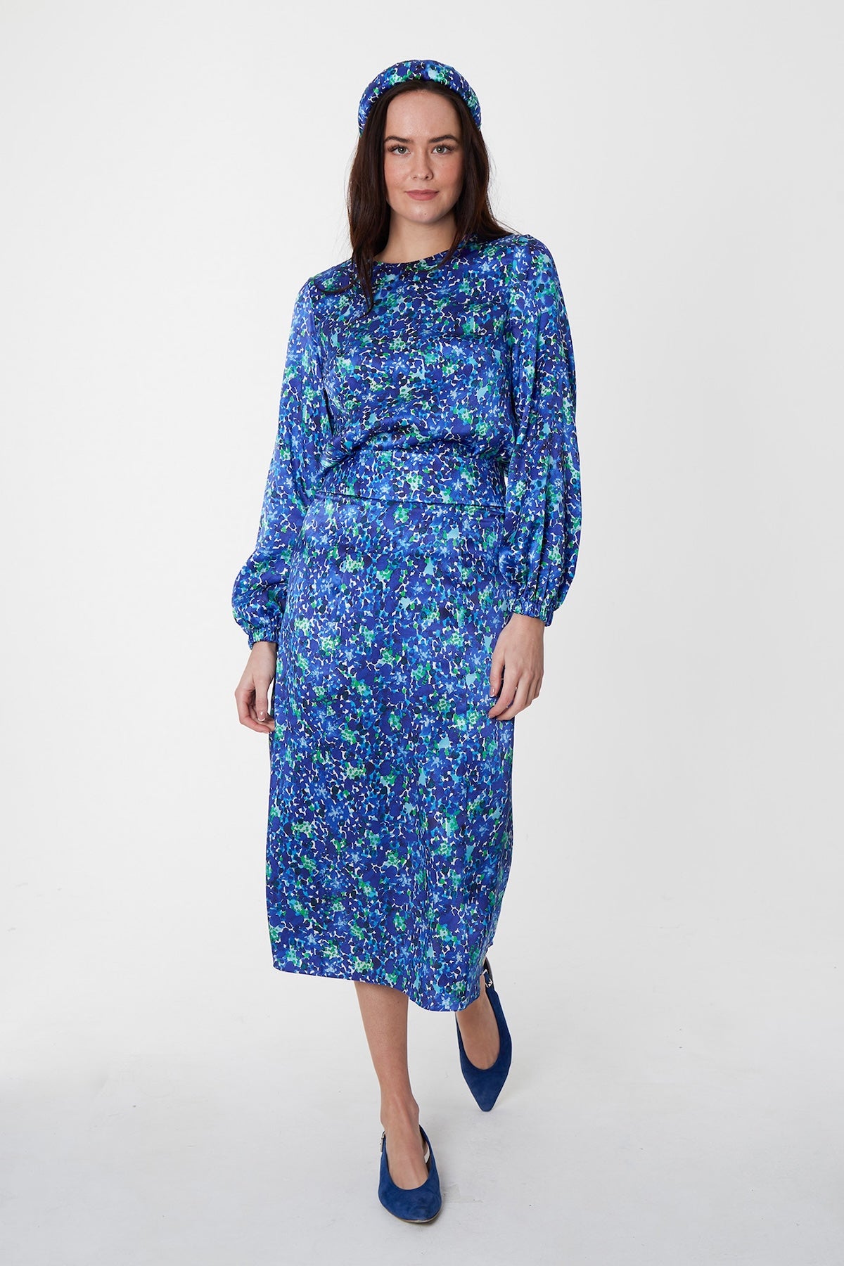 Model wearing blue floral silk printed Isabel Manns narrow a-line midi skirt with wide waistband, worn with matching long sleeve Emily Top and padded headband.