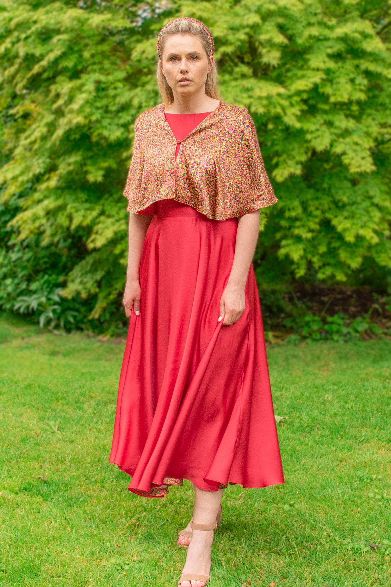 Long shot of model wearing reversible orange/pink dotted print silk satin shawl with pearl fastener at the chest, with matching padded headband and raspberry pink Victoria Gown.