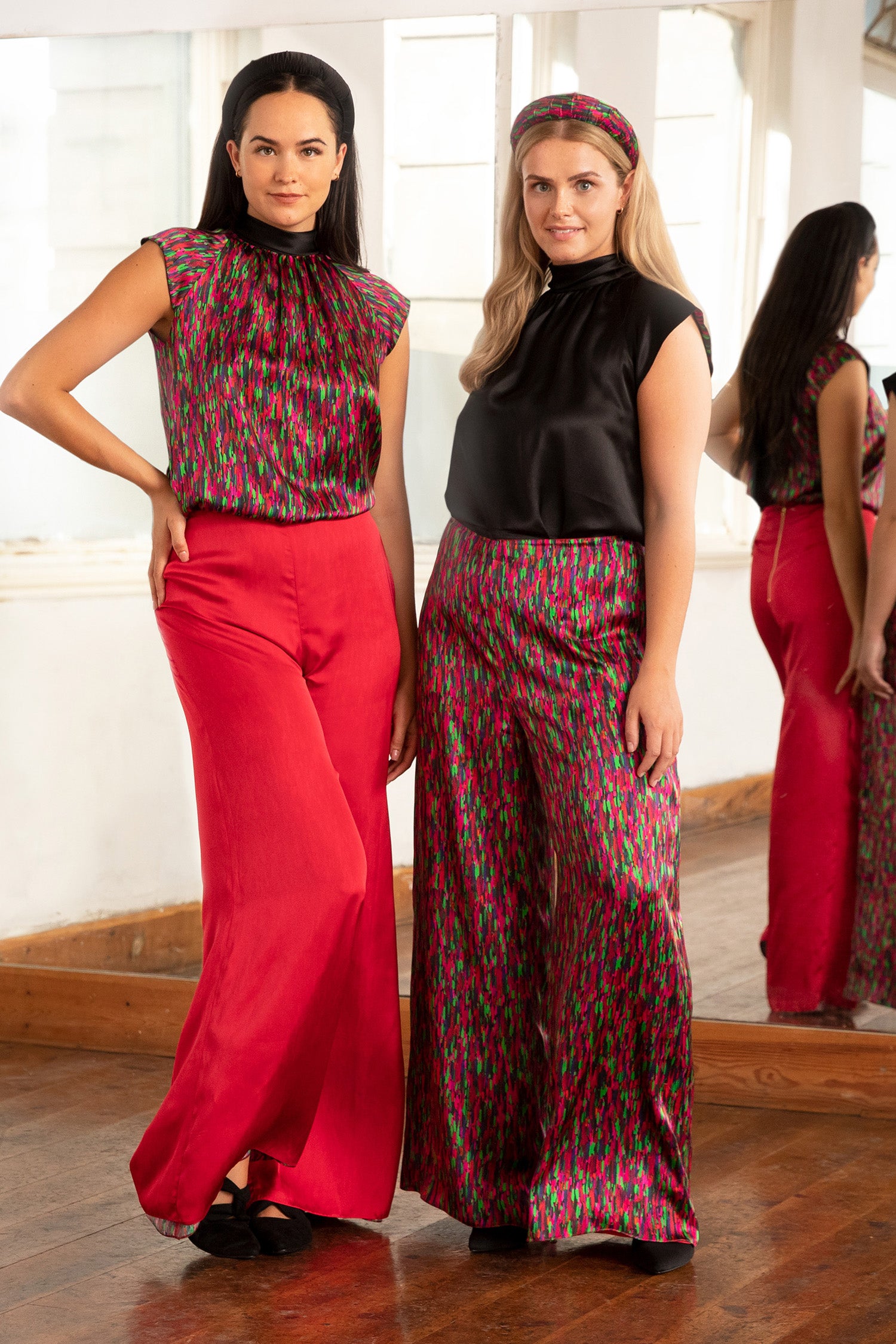 Two models wearing reversible high waisted silk satin palazzo pants in Raspberry Pink and matching halter neck cap sleeved Maya Top in pink dotted print, and another wearing reversible high waisted palazzo pants in dotted pink print and matching halter neck cap sleeved Maya Top in black.