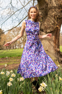 a purple printed a-line sleeveless dress worn by a model smiling and moving in a bed of tulips in a park