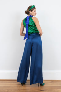 Back view of model wearing an Isabel Manns wide leg wrap around style silk viscose trousers in navy and contrasting vibrant green print lining, worn with matching halter neck Sophie Top in vibrant green print and padded headband.