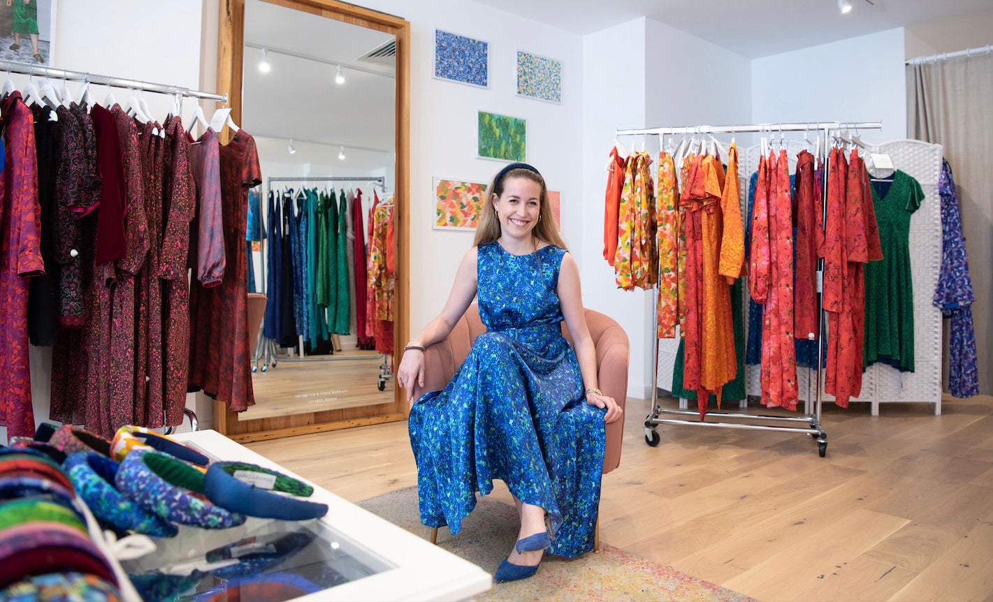An image of Isabel, the found of Isabel Manns smiling, sitting in her shop in a silk satin blue sleeveless long dress, showcasing the variety of products in her shop in the background