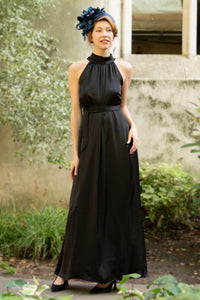 a black silk satin long halter neck style dress worn by a model showing the beautiful movement and waist cinched in with a tie belt, worn with a fascinator