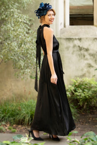 Side view of a black silk satin long halter neck style dress worn by a model and waist cinched in with a tie belt, worn with a fascinator