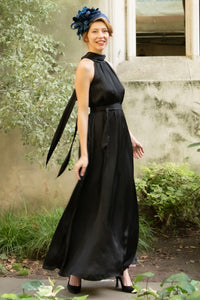 a black silk satin long halter neck style dress worn by a model and waist cinched in with a tie belt, worn with a fascinator