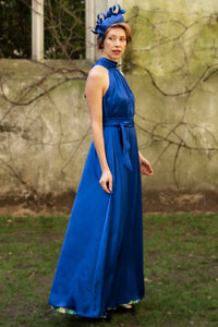 a cobalt silk satin long halter neck style dress worn by a model showing the beautiful movement and waist cinched in with a tie belt, worn with a fascinator