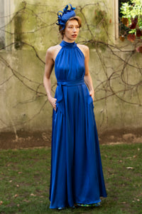 a cobalt silk satin long halter neck style dress worn by a model showing the beautiful movement and waist cinched in with a tie belt, worn with a fascinator