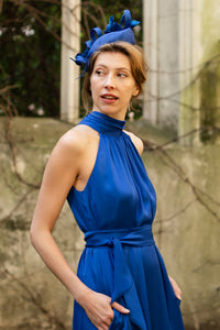 a detail shot of a cobalt silk satin halter neck dress with a tie belt, worn with a fascinator