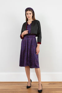 Model wearing reversible box shaped cropped black silk satin jacket, with gold bell fastening, worn with matching padded headband and purple print Valentina Dress.