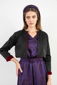 Model wearing reversible box shaped cropped black silk satin jacket, with gold bell fastening, worn with matching padded headband and purple print Valentina Dress.