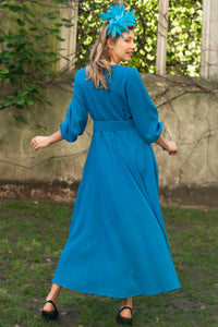 Back view of princess seam wool crepe dress in teal. Features 3/4 length elasticated sleeves, a rounded elegant neckline, and belt with buckle. Worn with fascinator.