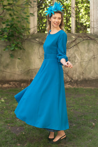 An Isabel Manns princess seam wool crepe dress in teal. Features 3/4 length elasticated sleeves, a rounded elegant neckline, and belt with buckle. Worn with fascinator.