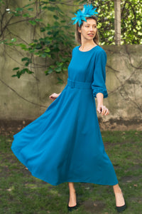 An Isabel Manns princess seam wool crepe dress in teal. Features 3/4 length elasticated sleeves, a rounded elegant neckline, and belt with buckle. Worn with fascinator.