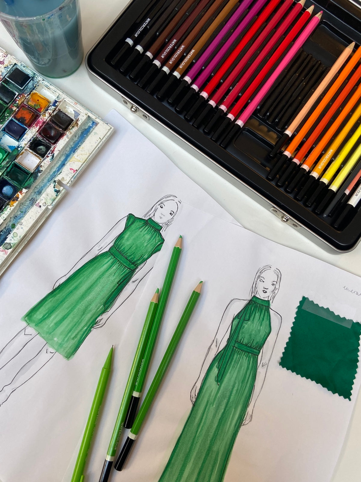 A sketch of two emerald green dress designs with pencils and paint surrounding it.