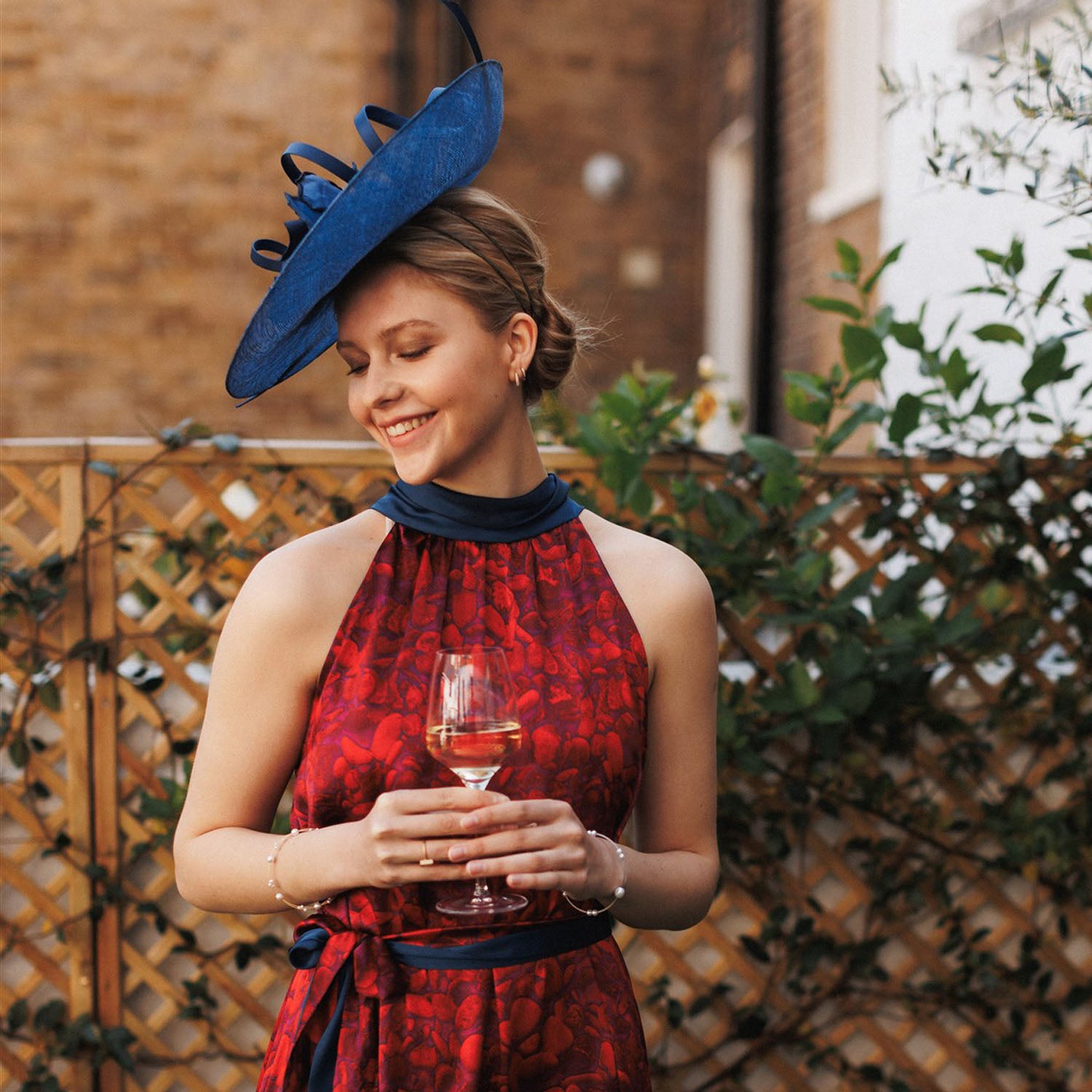 A beautiful lady smiling wearing an Isabel Manns red printed silk satin halter neck dress with matching blue hat in a garden drinking wine to showcase the elegance of our designs