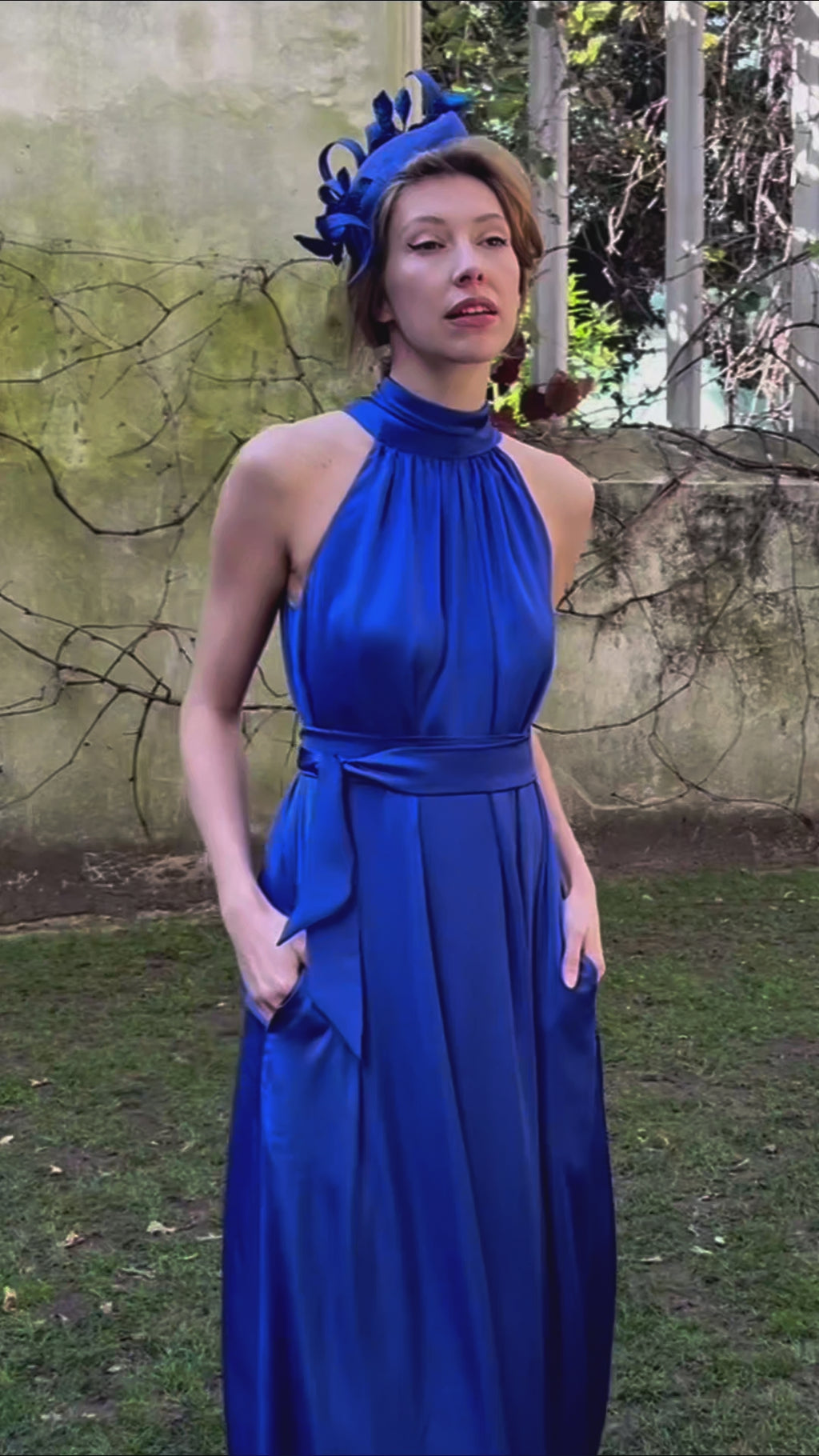 a cobalt silk satin long halter neck style dress worn by a model showing the beautiful movement and waist cinched in with a tie belt, worn with a fascinator