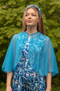 Close up view of Model wearing curved elbow length turquoise chiffon shawl with pearl fastener, worn with Penny Jumpsuit Serenity blue and matching padded headband.